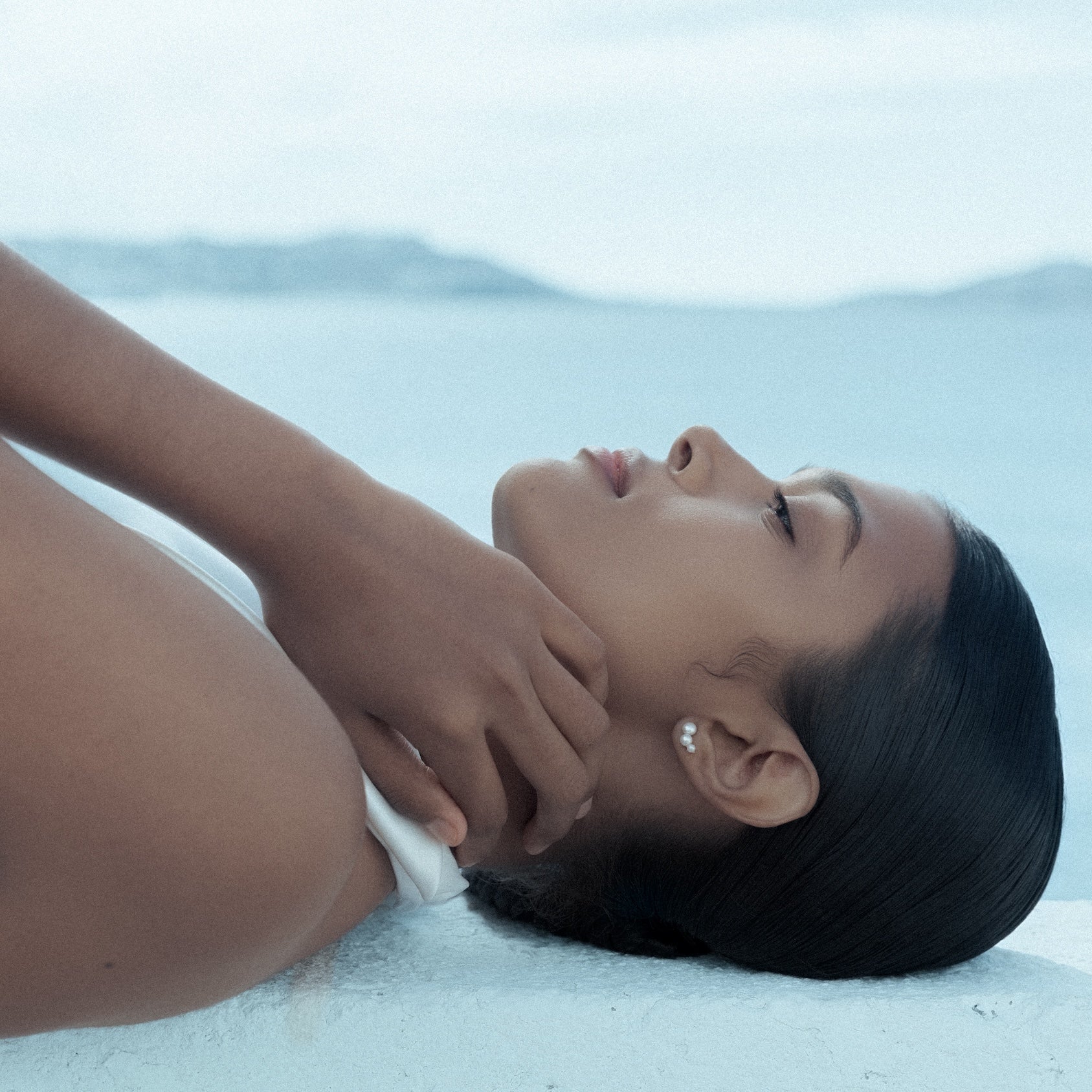 Woman lying on a white wall with a blurred sea background, wearing Calliadne earrings made of 3 freshwater pearls
